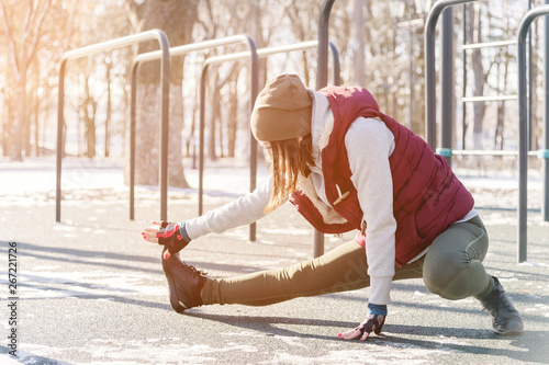 Sports girl in warm clothes on a sunny day does a warm-up on the outdoor sports field in the winter in spring or autumn during the cold season on the background of bars and horizontal bars