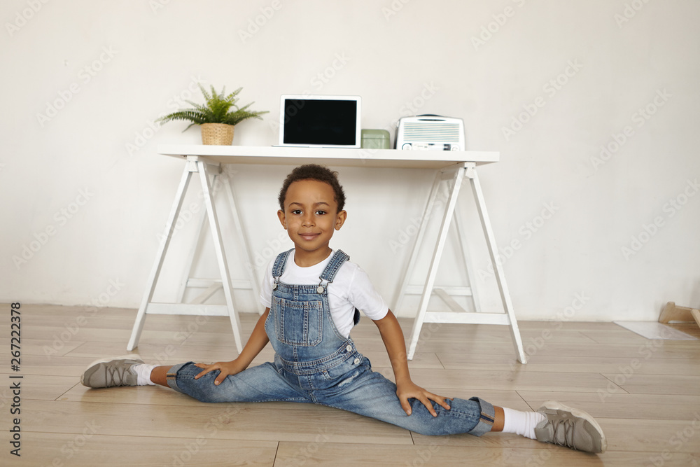 Horizontal image of sporty active flexible black African ten year old boy  in stylish clothes and sneakers doing side splits on floor with blank  copyspace laptop on white table in background Stock
