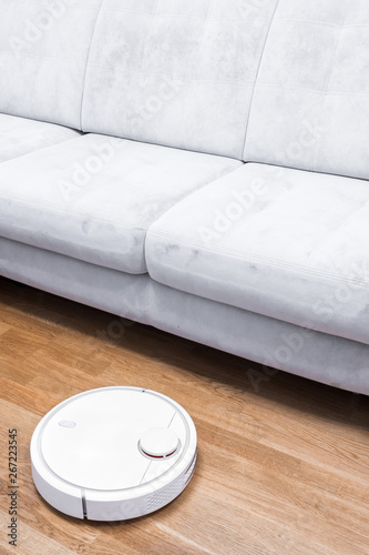 Robotic vacuum cleaner runs near sofa in room on laminate floor. Robot controlled by voice commands to direct cleaning. Modern smart cleaning technology housekeeping