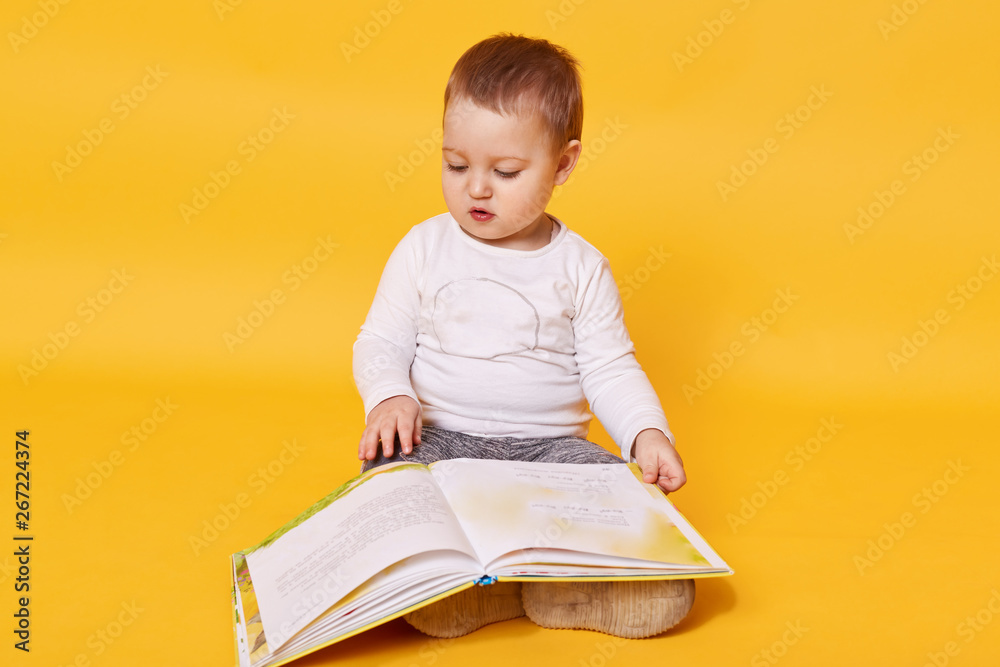 Toddler girl pretends to read book while sitting on floor, viewing pictures and turning pages, little girl looks concentrated, wearing casual white shirt, posing isolated over yellow background. Toddler girl pretends to read book while sitting on floor, viewing pictures and turning pages, little girl looks concentrated, wearing casual white shirt, posing isolated over yellow background.