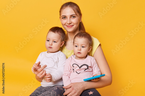 Charming affectionate mummy sitting with her cute little daughters on floor in yellow studio, females dressed casually, looks happy, children spending time with mom, sisters on mommy's knees.