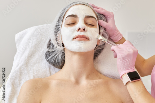 Spa and skin care concept. Young woman with facial mask in beauty salon, cosmetologist using special brush for applaying cream on client's face, female with closed eyes lying on comfortable couh.