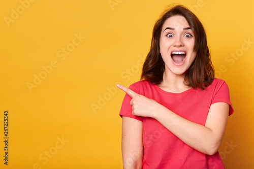 Indoor shot of positive attractive female with dark hair, dressed casually, points aside with index finger, has cheerful expression, shows something amazing at blank space, isolated on yellow wall.