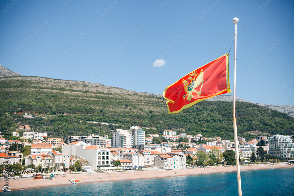 The national flag of Montenegro. In the background is Petrovac, the sea and the beach with architecture and buildings, hotels and houses.