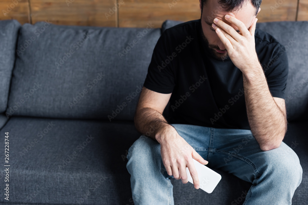 Fototapeta premium depressed man sitting on sofa with smartphone and holding hand on forehead