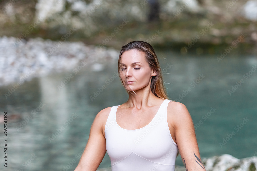 Inspired young beautiful woman and yoga trainer meditating sitting in a white suit in the lotus position with eyes closed in the wild nature against the background of the river and green hills