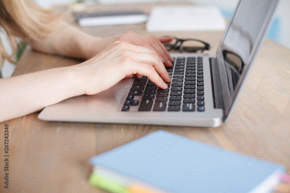 Side view of female hands typing on computer keyboard. Woman working on laptop sitting at work table. Glasses and Notes lying on wooden table. Laptop computer with blank screen Side view of female hands typing on computer keyboard. Woman working on laptop sitting at work table. Glasses and Notes lying on wooden table. Laptop computer with blank screen
