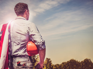 Handsome man with tools, holding an American Flag, standing against a background of green trees and the rays of the setting sun. View from the back, close-up. Concept of work and employment