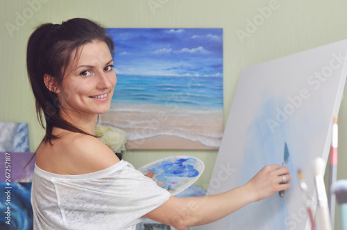 A beautiful woman artist paints a picture of blue acrylic paints on a large white canvas at her home against the background of her painting of the sea.