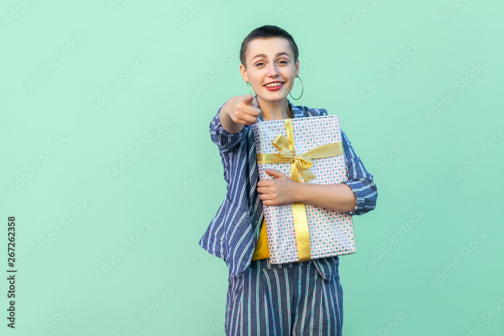 Winner are you! Portrait of happy beautiful with short hair young woman in striped suit standing, hugging present box and pointing finger to camera. Indoor, isolated, studio shot, green background Winner are you! Portrait of happy beautiful with short hair young woman in striped suit standing, hugging present box and pointing finger to camera. Indoor, isolated, studio shot, green background