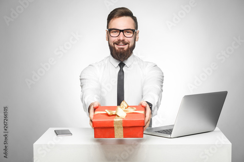 Take present! Portrait of happy young boss in in white shirt and black tie are sitting in office and giving you a red box, congratulate you with holiday. Indoor, isolated, studio shot, gray background