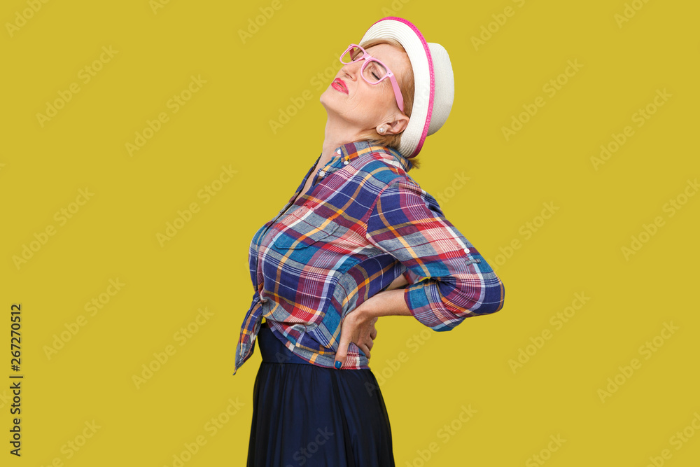 Back, spine or kidney pain. Profile side view portrait of modern stylish mature woman in casual style with hat standing and holding her painful back. indoor studio shot isolated on yellow background. Back, spine or kidney pain. Profile side view portrait of modern stylish mature woman in casual style with hat standing and holding her painful back. indoor studio shot isolated on yellow background.
