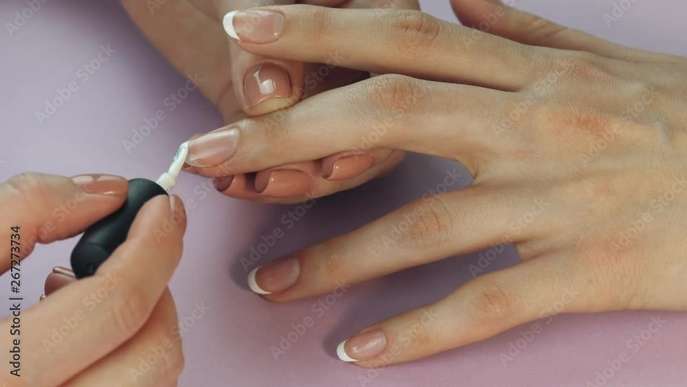 the manicurist paints the client's nails with nail polish on a lite purple background Painting nails. Female hands with beautiful french manicure the manicurist paints the client's nails with nail polish on a lite purple background Painting nails. Female hands with beautiful french manicure