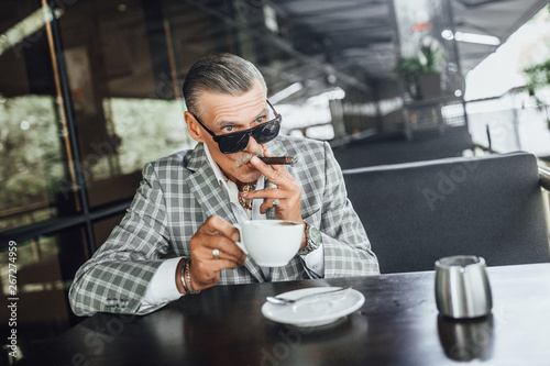 Old senior man holding a cigarette sitting on a balcony and drinking latte