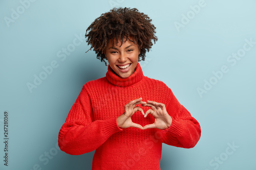 Portrait of lovely female model makes heart gesture, says be my valentine, demonstrates love sign, has glad expression, wears warm red jumper, isolated against blue background. Body language