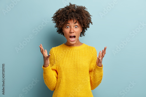 Emotive stunned Afro American female gestures while screams loudly, scared of horror film, wears bright knitted yellow jumper, isolated against blue background. Human emotions and feelings concept