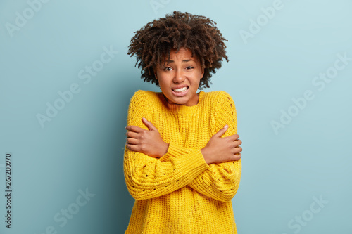 Scared fearful dark skinned woman trembles as afraids something awful, crosses hands over chest, clenches teeth, has Afro hairstyle, wears loose yellow sweater, models against blue background