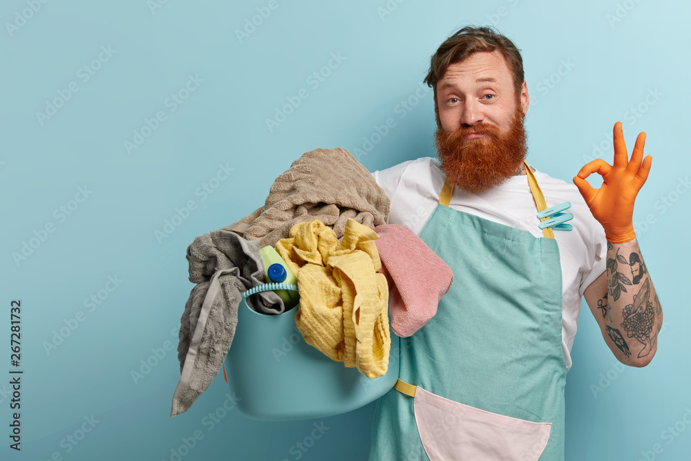 Satisfied bearded red haired man makes okay gesture, shows approval sign, says everything is under control, busy with daily routines, holds basin with dirty laundry, isolated on blue background - obrazy, fototapety, plakaty 