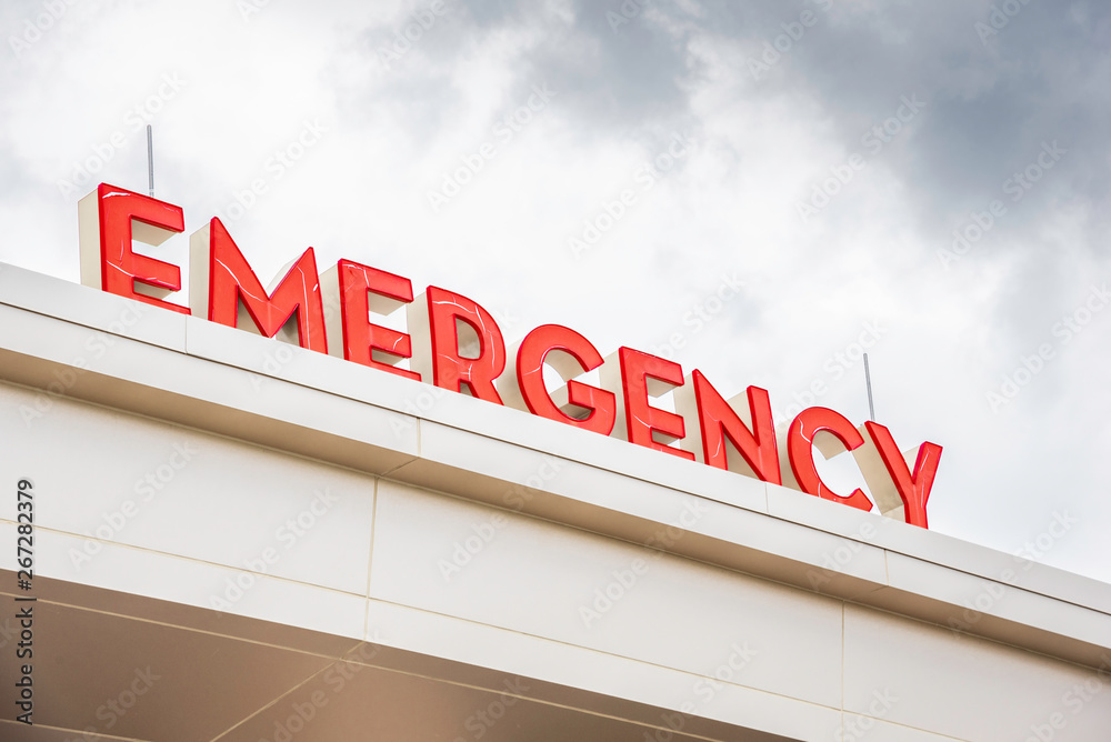 A prominent red 3D all-caps lighted emergency directional sign and marker perched on the awning and canopy of the main hospital entrance.