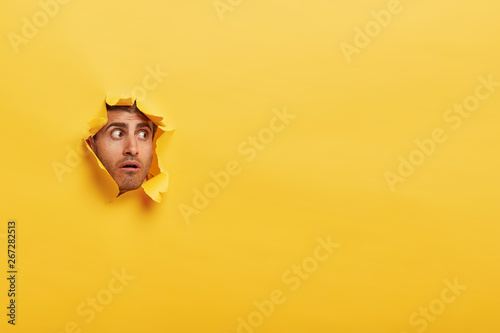 Astonished unshaven man has stupefied expression, peeks through torn wall paper, has widely opened eyes, stares through hole in yellow background. Human facial expressions and emotions concept photo