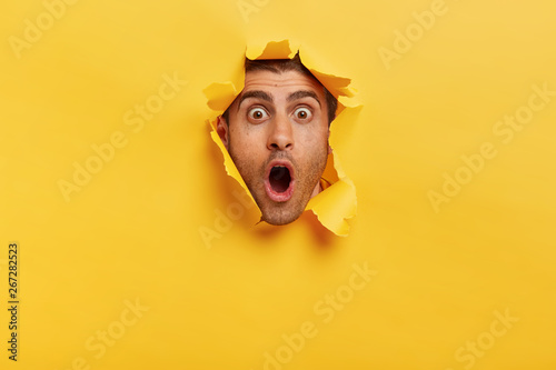 Stupefied young man with bristle keeps jaw dropped from surprisement, has popped eyes, stares through yellow torn paper, impressed by shocking rumors. People, human emotions and reaction concept photo