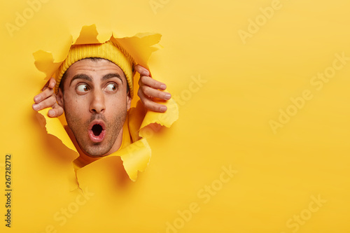 Surprised male face through paper hole. Emotional astonished young man wears yellow headgear, makes slot in background with hands, keeps mouth wide opened. Copy space to insert yout text or slogan photo