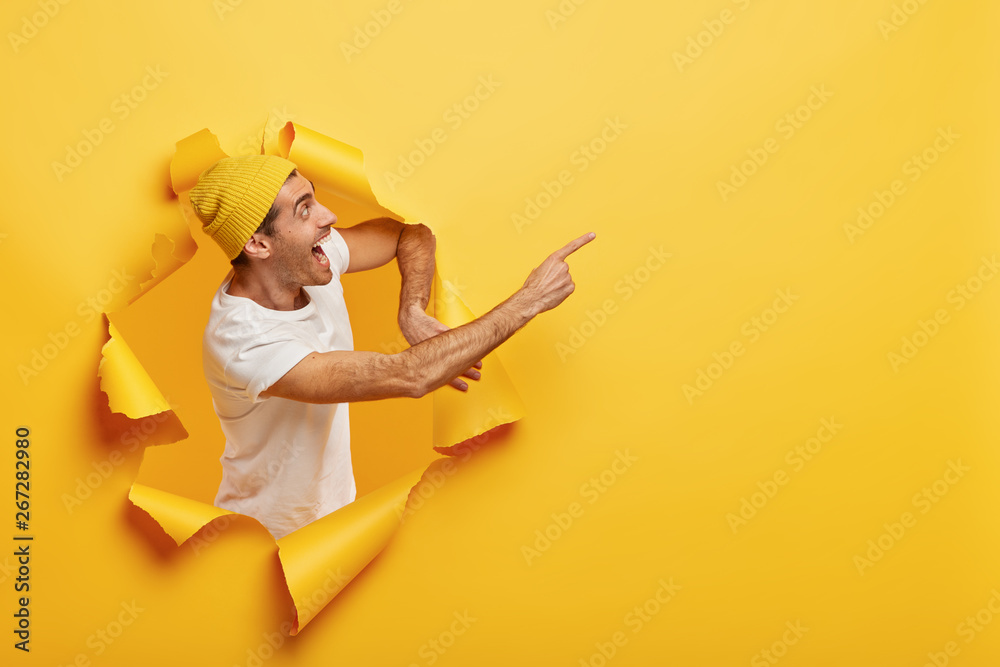 Photo of overjoyed hipster looks gladfully away, points fore finger on blank space, shouts from joy, stands through torn paper hole, wears stylish yellow hat and casual white t shirt. Promotion Photo of overjoyed hipster looks gladfully away, points fore finger on blank space, shouts from joy, stands through torn paper hole, wears stylish yellow hat and casual white t shirt. Promotion