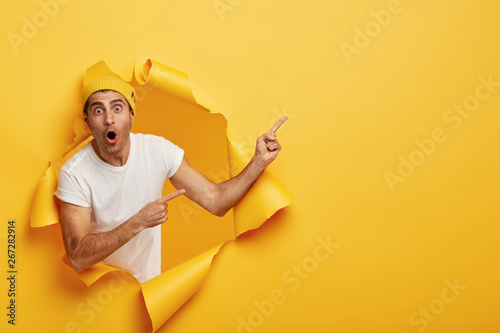 Horizontal shot of surprised young Caucasian man in white t shirt and yellow headgear, has widely opened mouth, indicates at copy space, models in torn paper background. Wow, just look there photo
