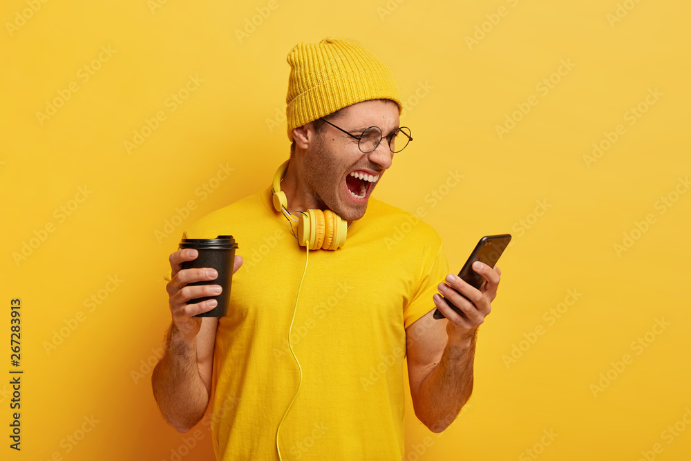 Irritated guy looks angrily at smart phone, receieves unpleasant message, screams negatively, wears yellow stylish outfit, round spectacles drinks coffee to go, stands in studio. Monochrome shot