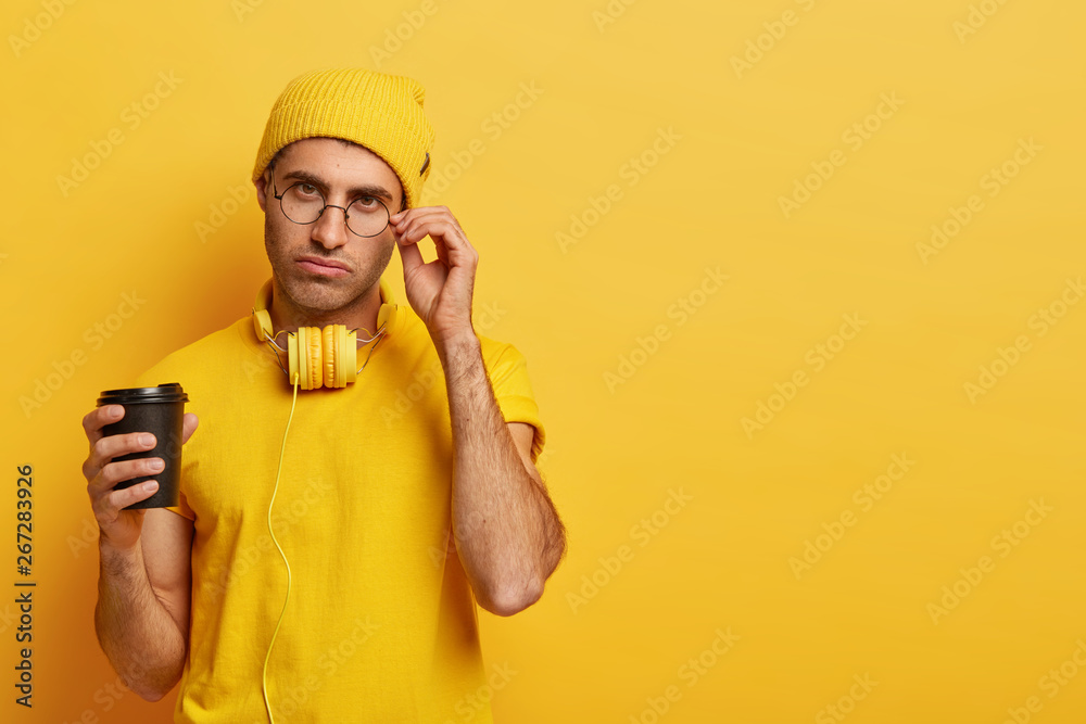 Serious male student keeps hand on rim of glasses, has tired expression, tries to refresh himself with strong coffee and music, wears yellow hat and t shirt, stands indoor with free space aside Serious male student keeps hand on rim of glasses, has tired expression, tries to refresh himself with strong coffee and music, wears yellow hat and t shirt, stands indoor with free space aside