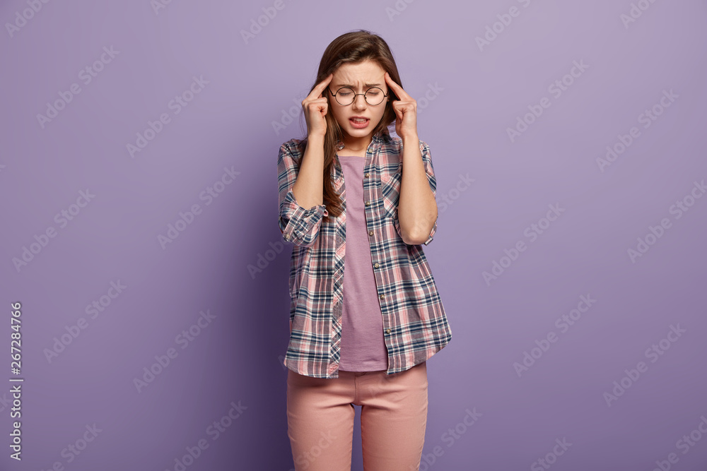 Stressful overworked woman keeps fingers on temples, suffers from headache or migraine, clenches teeth and frowns face from pain, has eyes closed, wears transparent glasses for vision correction Stressful overworked woman keeps fingers on temples, suffers from headache or migraine, clenches teeth and frowns face from pain, has eyes closed, wears transparent glasses for vision correction