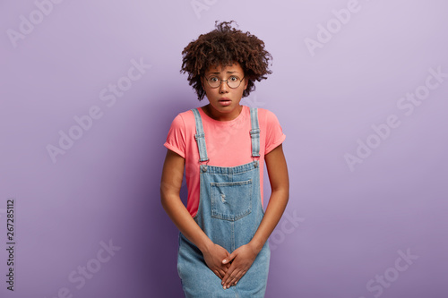 Photo Dissatisfied girl keeps hands on crotch, presses lower abdomen, needs toilet badly, has syndrome of cystitis, wears spectacles, pink t shirt and denim sarafan, isolated on purple wall