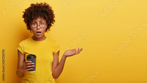 Horizontal studio shot of indignant woman raises hand, looks with puzzled expression, has talk with interlocutor during coffee break, holds paper cup, frustrated by latest bad news. Monochrome photo