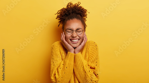 Quadro su tela Portrait of cheerful woman feels entertained and amused, watches funny show, touches cheeks with both hands, shows white teeth, keeps eyes closed, wears yellow jumper, stands indoor
