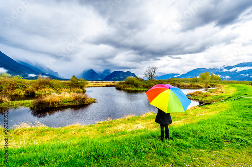 Woman with a rainbow colored Umbrella under dark rain clouds on a cold spring day at the lagoons of Pitt-Addington Marsh in Pitt Polder near Maple Ridge in British Columbia  Canada 