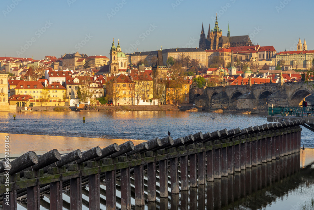 Outdoor sunny view of Charles Bridge, Prague Castle, St. Vitus Cathedral,  buildings on riverside, and wooden piles barrier stand on Vltava River in Prague, Czech Republic against golden sunrise sky.