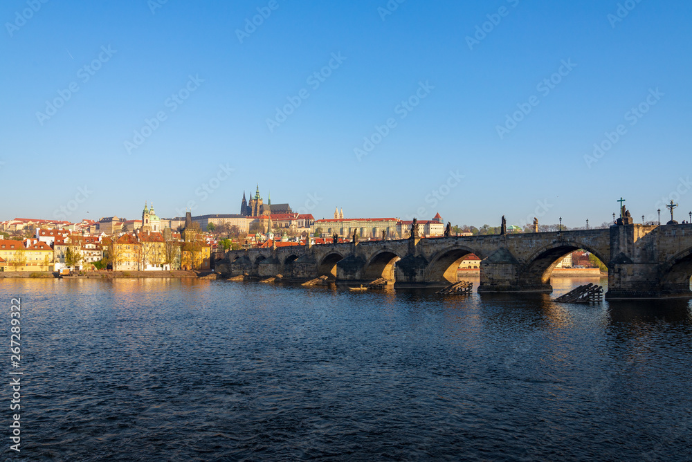 Outdoor sunny view of Charles Bridge, Prague Castle, St. Vitus Cathedral and riverside of Vltava River in Prague, Czech Republic in the morning with blue sunrise sky and daybreak atmosphere.