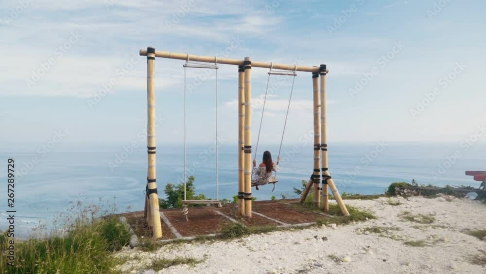 Backside view of young woman swings on swing with beautiful view on ocean, slow motion Backside view of young woman swings on swing with beautiful view on ocean, slow motion