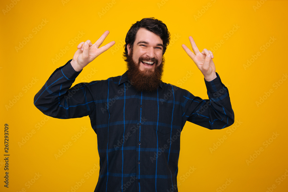 A cheerful man with beard and toothy smile, doing peace gesture over yellow background A cheerful man with beard and toothy smile, doing peace gesture over yellow background