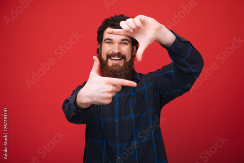 Photo of handome bearded man standing over red background and doing a photo frame with fingers, taking a photo photo