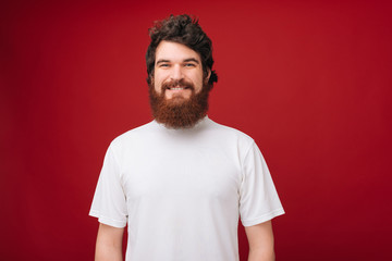 Portait of cheerful bearded guy in white tshirt smiling and looking at camera over red background