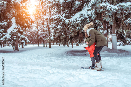 A young mother supports a young son, a boy of 3 years. In winter, outside in park, background is snow drifts of Christmas tree. Free space. Children's skis, skiing lessons, first steps, child care.