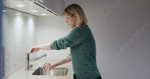 Young blonde woman filling a glass of water and drinking in the kitchen