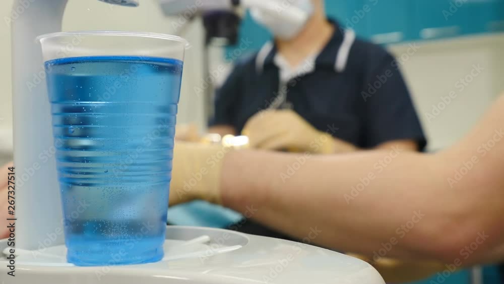 Blurred dental procedure in modern dentist office. Space for text. Plastis cup with blue liquid on foreground is in focus. Male dentist examining patient. 4k Blurred dental procedure in modern dentist office. Space for text. Plastis cup with blue liquid on foreground is in focus. Male dentist examining patient. 4k