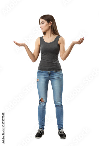 Young woman in gray top and blue jeans standing with arms bent and raised at sides looking at one of her palms facing up isolated on white background.