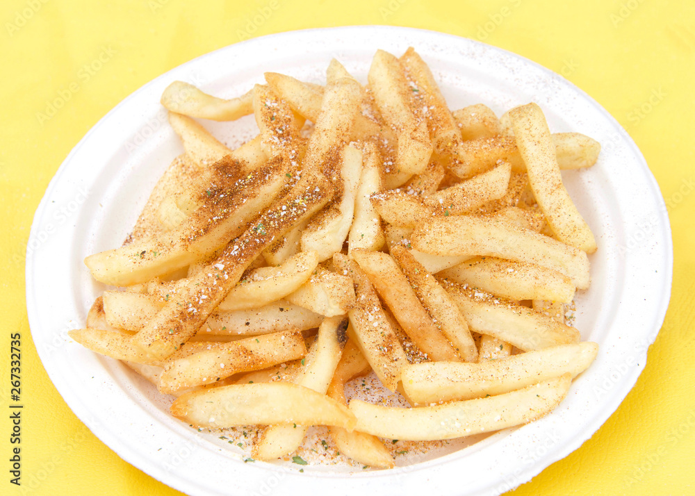 White paper plate with seasoned french fries on yellow table cloth. Popular street fair food. White paper plate with seasoned french fries on yellow table cloth. Popular street fair food.