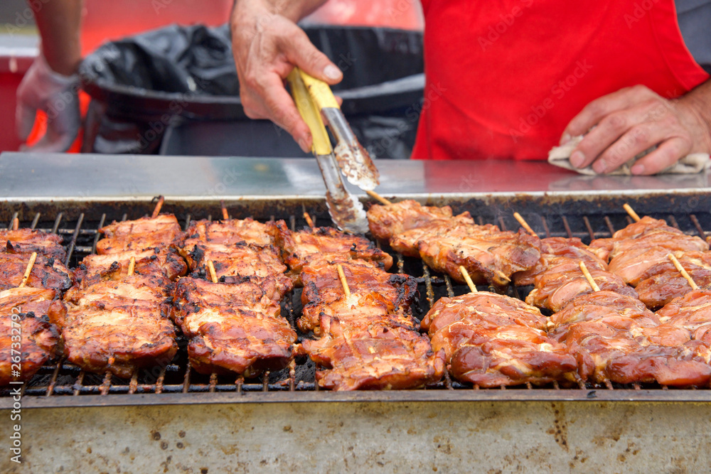 Close up on chicken filets skewered and grilling on outdoor grill, person with tongs wearing red apron flipping the meat. Popular street fair food.