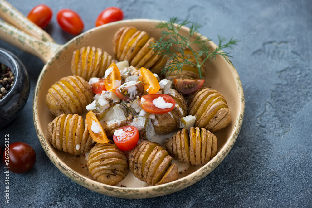 Close-up of hasselback potatoes with tartare sauce and cherry tomatoes in a serving pan, studio shot over blue stone surface Close-up of hasselback potatoes with tartare sauce and cherry tomatoes in a serving pan, studio shot over blue stone surface