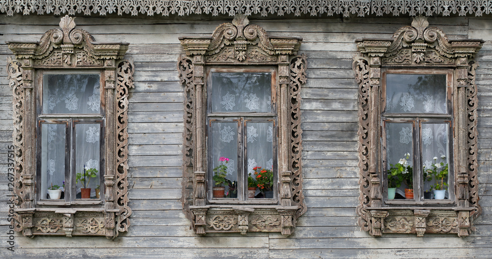 Three village windows with carved platbands and a cornice of the national Russian style against the background of aged faded wooden boards Three village windows with carved platbands and a cornice of the national Russian style against the background of aged faded wooden boards