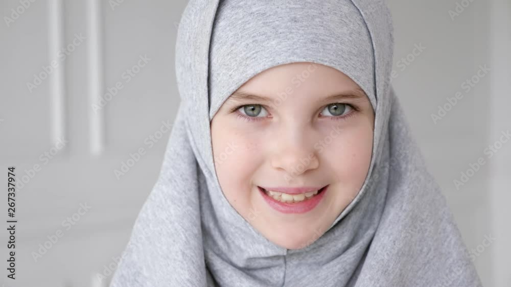 Portrait of young attractive muslim 9-years girl wearing grey hijab and traditional dress is rising her eyes, looking at the camera and smiling on light background. Portrait of young attractive muslim 9-years girl wearing grey hijab and traditional dress is rising her eyes, looking at the camera and smiling on light background.